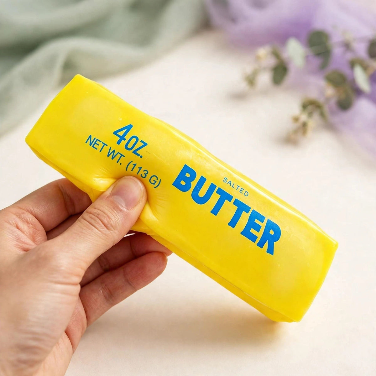 Butter Stick Squishy — soft yellow butter stick squeeze toy being squeezed by hand on a cream background